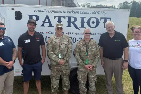 Group of six people standing in front of a Patriot trailer donated to Jackson County JROTC outdoors on grassy hill.