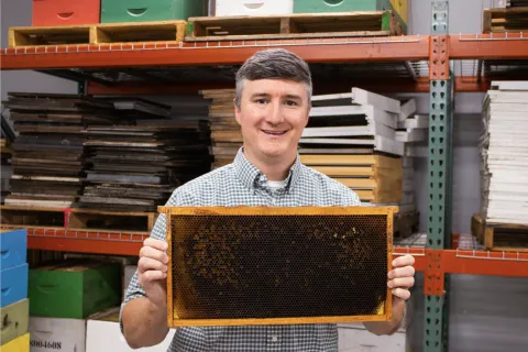 Smiling man holding a honeycomb frame in a bee farm with shelves and boxes in the background.