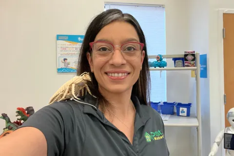 Woman with glasses smiling in a lab with educational posters, toy dinosaurs, and a small robot on shelves