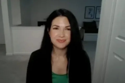 Smiling woman with long dark hair in a black top sitting indoors in a modern, light-colored room.