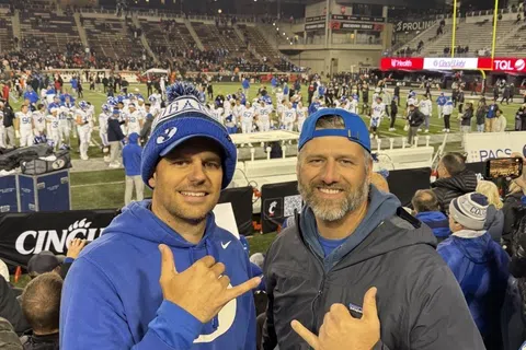 Two men in sports apparel making shaka signs at a crowded football stadium during a night game.