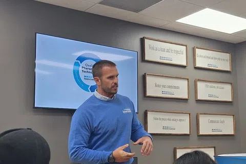 Man in blue sweater presenting to a small group in a conference room with laptops and visual aids on the walls.