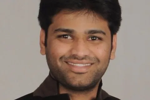Smiling young man with dark hair wearing a black jacket and orange shirt against a plain background