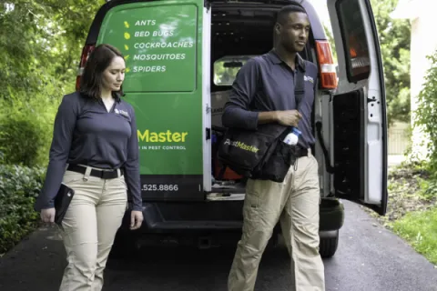 Two pest control professionals walking from a van with a list of pests serviced and equipment in hand