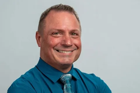Smiling man in a blue dress shirt and patterned tie with arms crossed against a light gray background
