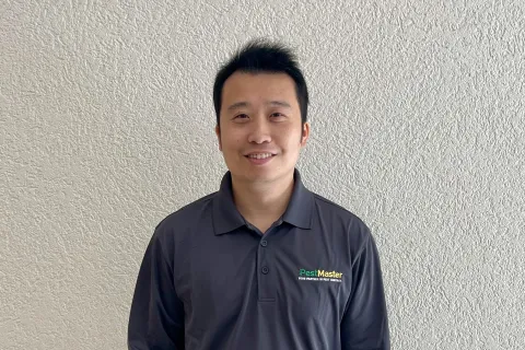 Smiling man standing against textured wall wearing dark long sleeve shirt with PestMaster logo and black jeans.