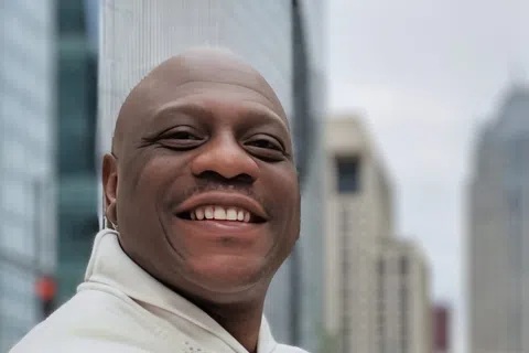 Smiling man in white sweater with arms crossed standing outdoors in an urban cityscape with tall buildings.