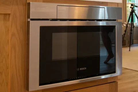 Stainless steel Bosch microwave built into wooden kitchen cabinetry with white countertop.