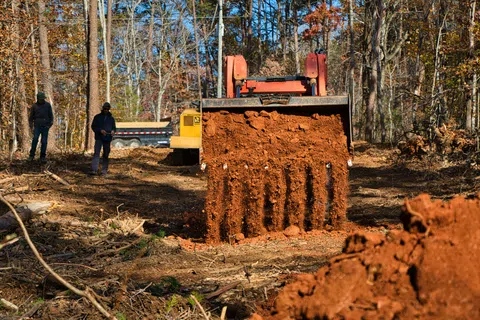 Bulldozer spreading rich red soil on a cleared forest site with two workers observing nearby under clear sky.