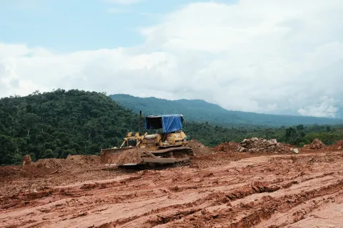 Bulldozer with a blue cover working on muddy terrain near a forested mountain under cloudy sky.