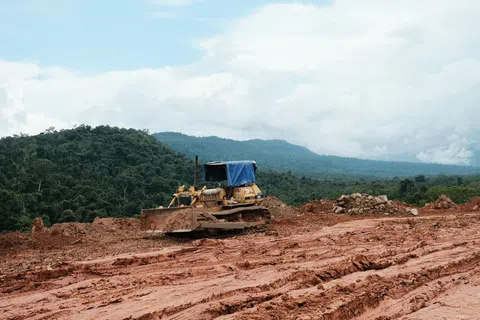 Bulldozer working on clearing a muddy construction site near forested hills under a cloudy sky.