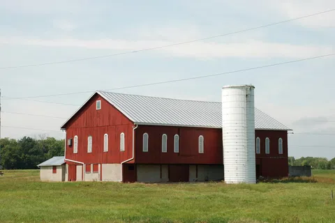 Red barn with white silo in green field under a partly cloudy blue sky during daytime.