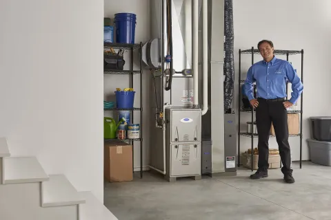 Technician stands in a clean basement near a modern HVAC furnace and shelving with containers and tools.