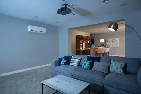 Cozy living room with gray sectional sofa, colorful pillows, coffee table, wall-mounted AC, and projector.