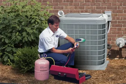 Technician servicing an outdoor Carrier air conditioning unit with tools and refrigerant tank near brick wall.