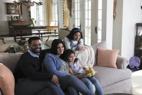 Smiling family of four sitting on a gray couch in a bright, cozy living room with large windows and soft decor.