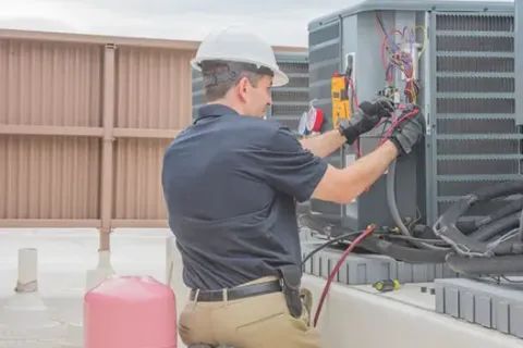 Technician in hard hat working on rooftop HVAC unit wiring with multimeter and gloves under daylight.