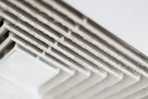 Close-up of a dusty white air vent with accumulated dirt on the metal slats indoors.