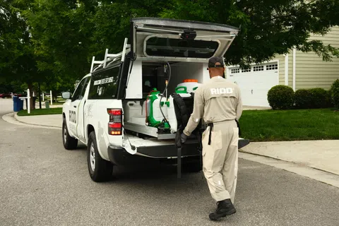 Pest control worker loading equipment into a white RIDO service truck parked in a residential neighborhood.