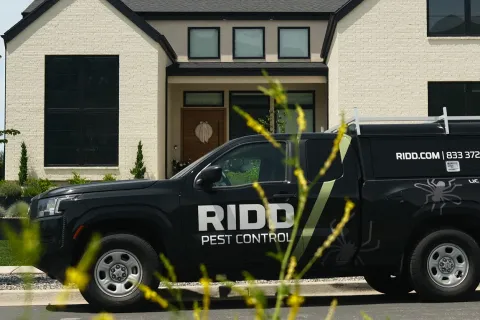 Black RIDDO Pest Control truck parked outside a modern suburban house with technician standing nearby.