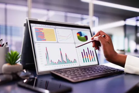 Person using stylus to analyze colorful charts and graphs on a laptop screen in a modern office.