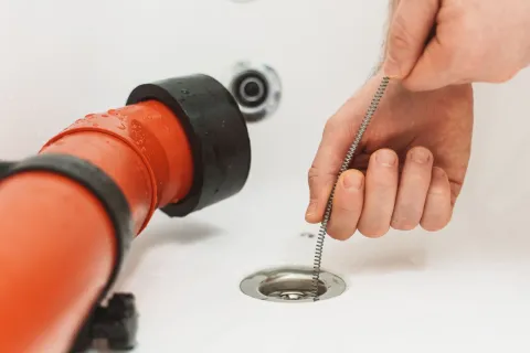 A person unclogging a sink with a plumbing snake near a red drainage pipe.