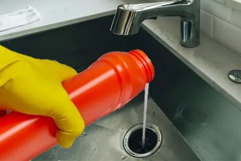 Person wearing yellow gloves pouring drain cleaner from an orange bottle into a kitchen sink drain below a chrome faucet.