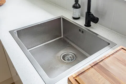 Modern kitchen sink with black faucet, wooden cutting board on white countertop near window with fruit bowl.