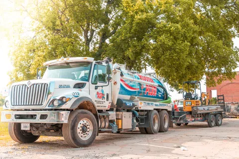 Rooder Plus septic service truck with tank and small excavator on trailer parked under green trees on sunny day.