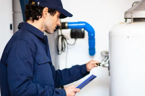 A technician inspecting a water heater, holding a clipboard and adjusting a valve.