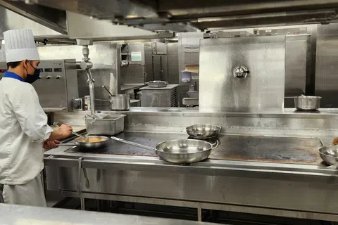 Chef wearing white uniform and hat cooking in a professional stainless steel kitchen with pans on stove.