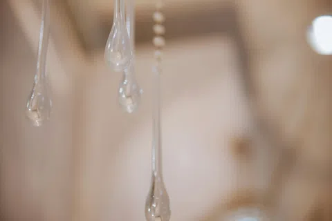 Close-up of several elongated glass droplets hanging from strings against a blurred beige background.