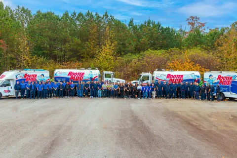 Large Rooter Plus! plumbing team standing in front of five service trucks outdoors with trees in fall colors behind them.