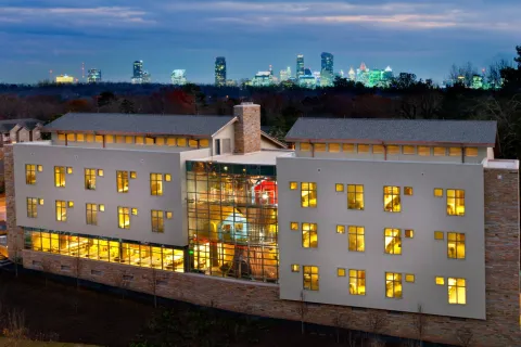 Modern illuminated building with large glass facade and city skyline at dusk with light trails on nearby road.