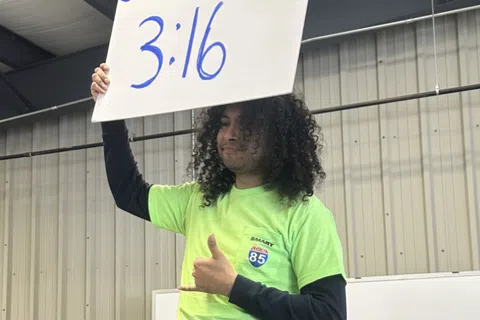 Man with curly hair wearing a championship belt and neon green shirt holds a sign reading Screws 3:16 indoors.