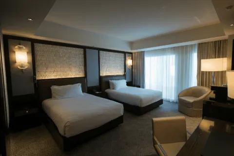 Modern hotel room with two beds, upholstered chair, desk, soft lighting, and large window with curtains.