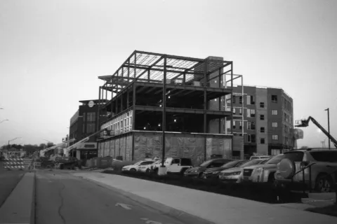 Black and white photo of a multi-story building under construction with parked cars along the street.