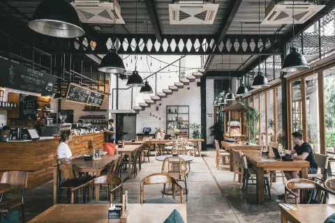 Spacious modern cafe interior with wooden furniture, industrial lighting, and large windows letting in natural light.