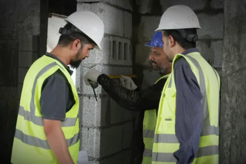 Three construction workers in helmets and reflective vests measuring a concrete block wall inside a building