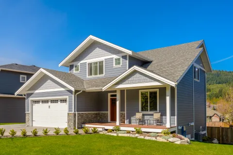 Modern two-story gray house with stone accents, front porch, garage, and green lawn under clear blue sky.