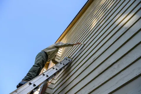 A professional painter on a ladder applying a fresh coat of beige paint to horizontal siding, illustrating how choosing the right exterior colors can increase home value.