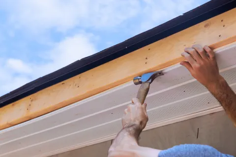 Close-up of a person using a hammer to install wooden fascia board under a roof against blue sky.