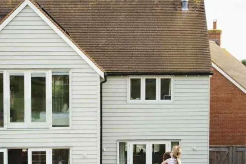 Father carrying daughter on his back walking towards a modern suburban house with large windows and green lawn