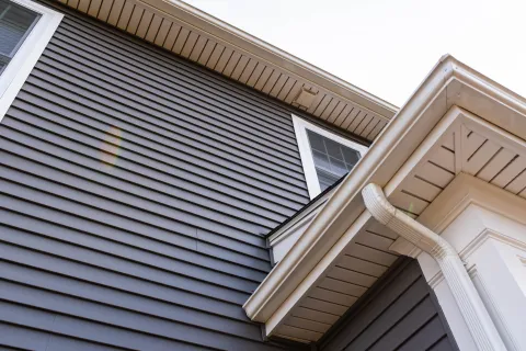 Close-up of a house exterior showing gray vinyl siding, white trim, gutter, and window under a clear sky.