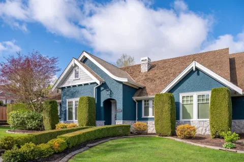 Blue one-story house with brown roof, white trim, and manicured green lawn with shaped bushes under a blue sky.