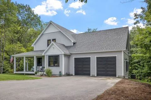 Modern two-story white house with black garage doors, covered porch, and surrounding green trees under blue sky.