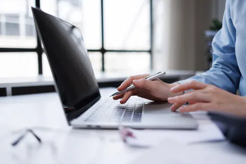 Person working on a laptop with one hand holding a pen and the other typing on the keyboard in bright office light.