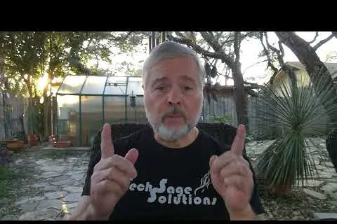 Middle-aged man with gray beard gestures with both hands outdoors near greenhouse and plants at sunset.