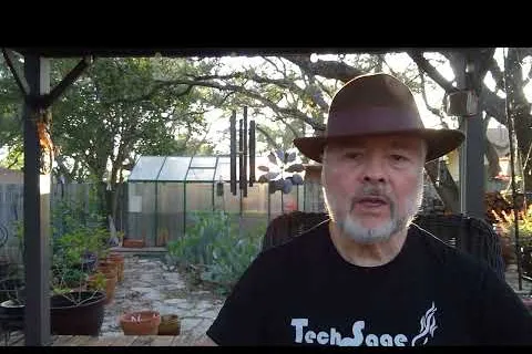 Older man wearing brown hat and TechSage shirt sitting outdoors in garden with greenhouse behind him.