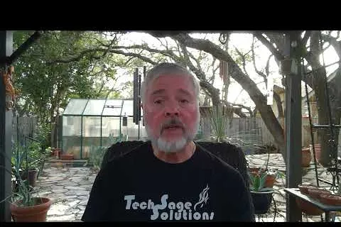 Bearded man wearing a black Tech Sage Solutions shirt sitting outdoors with plants and greenhouse in background